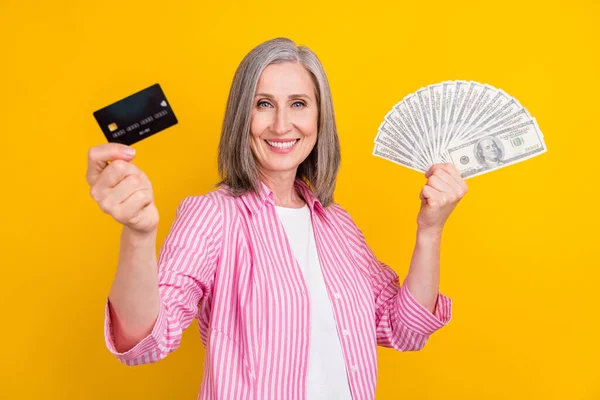 Foto di donna anziana felice sorriso positivo tenere i soldi della carta di credito dollari in contanti isolato su sfondo di colore giallo — Foto Stock