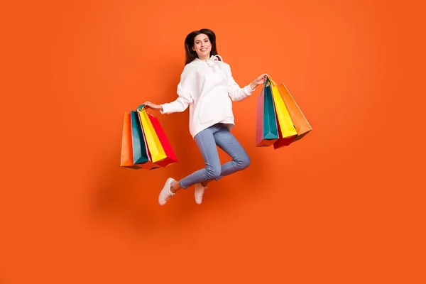 Vista completa del tamaño del cuerpo de la atractiva chica alegre saltando llevando bolsas de salida ofrecen aislado sobre fondo de color naranja brillante — Foto de Stock