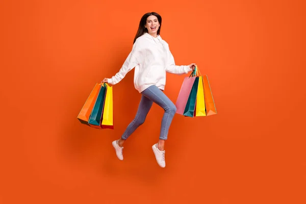 Full length body size view of pretty cheerful girl jumping carrying bags isolated over bright orange color background — Stock Photo, Image