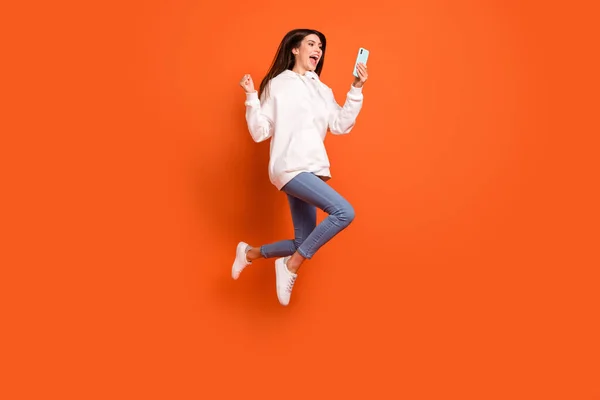 Tamanho do corpo de comprimento total vista de menina alegre sorte muito na moda saltando usando dispositivo ganhar isolado sobre fundo de cor laranja brilhante — Fotografia de Stock