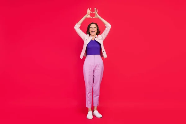 Foto av romantisk gammal dam höja händerna visa hjärta gest bära rosa jacka isolerad röd färg bakgrund — Stockfoto