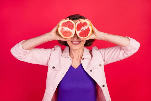 Foto von Rentnerin Dame halten zwei Grapefruit Scheibe Deckel Augen tragen rosa Jacke isoliert rote Farbe Hintergrund — Stockfoto