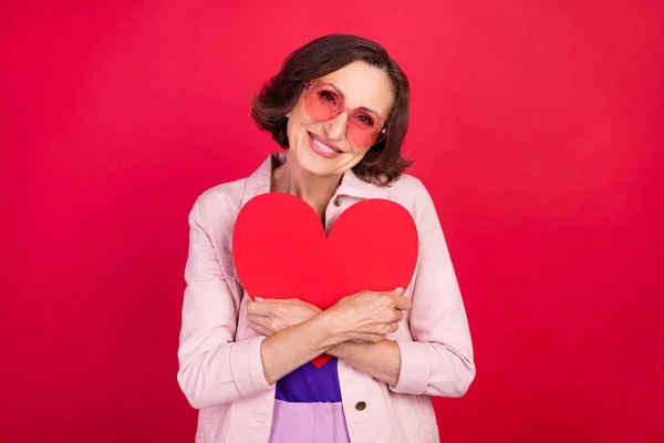 Fénykép a romantikus idős hölgy ölelés szív kártya sugárzó mosoly viselet napszemüveg rózsaszín kabát elszigetelt piros szín háttér — Stock Fotó
