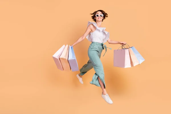 Foto van lieve schattige vrouw gekleed gestreepte top donkere bril holding tassen springen lopen geïsoleerde beige kleur achtergrond — Stockfoto