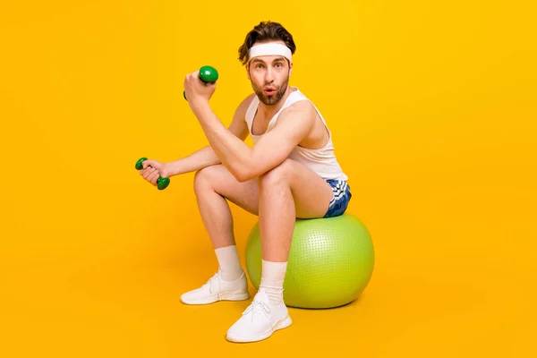 Retrato de chico musculoso atractivo sorprendido sentado en la bola levantamiento de peso trabajar aislado sobre fondo de color amarillo brillante —  Fotos de Stock