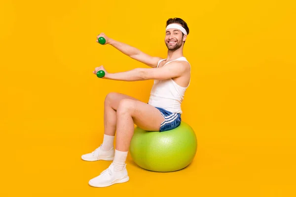 Ganzkörpergröße Profil Seitenansicht des sportlichen fröhlichen Kerl sitzt auf Ball heben Gewicht isoliert über helle gelbe Farbe Hintergrund — Stockfoto