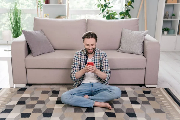 魅力的な焦点を当てた陽気な男の肖像上に座ってカーペットを使用してデバイスsmmアプリ5g家にいます。 — ストック写真