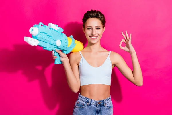 Porträt von attraktiven fröhlichen Mädchen mit Wasserpistole zeigt ok-Zeichen isoliert über hell rosa Magenta Farbe Hintergrund — Stockfoto