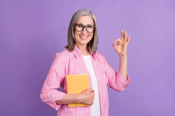 Foto de mulher madura sorriso feliz mostrar tudo bem bem anúncio sinal segure materiais do livro isolado sobre fundo de cor roxa — Fotografia de Stock