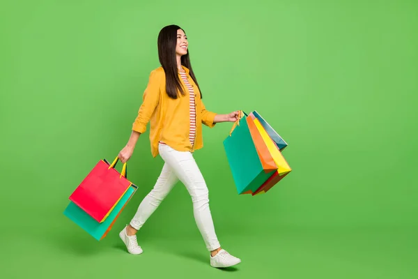 Ganzkörper-Profil-Foto von coolen brünetten Millennial Lady gehen mit Taschen tragen Hemdhosen isoliert auf grünem Hintergrund — Stockfoto
