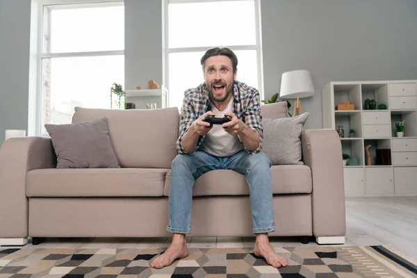 Foto de dulce divertido joven usar camisa a cuadros sofá sentado celebración playstation sonriendo en el interior habitación de la casa — Foto de Stock