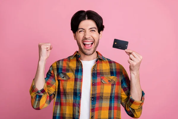 Foto de bastante afortunado joven caballero usar traje de cuadros sosteniendo tarjeta de crédito puño ascendente sonriendo aislado color rosa fondo — Foto de Stock