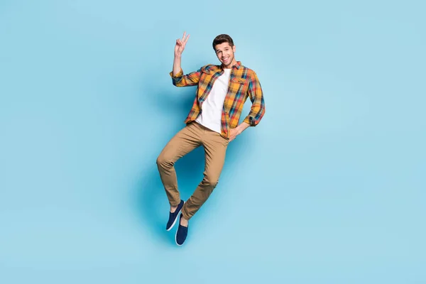 Foto de joven caballero atractivo funky usar camisa a cuadros saltando alto mostrando v-signo sonriente aislado color azul fondo —  Fotos de Stock