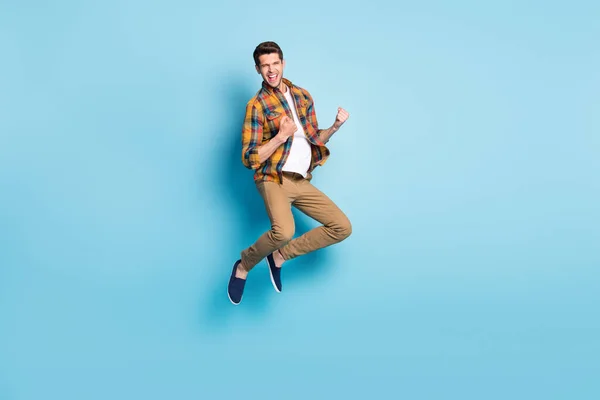 Фото беззаботного счастливчика в клетчатой рубашке прыгающего высоко поднимающегося кулака улыбающегося изолированного синего цвета фона — стоковое фото