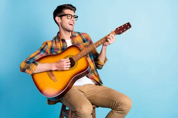 Foto de encantador engraçado jovem cavalheiro usar camisa xadrez cantando tocar guitarra espaço vazio sorrindo isolado azul cor de fundo — Fotografia de Stock