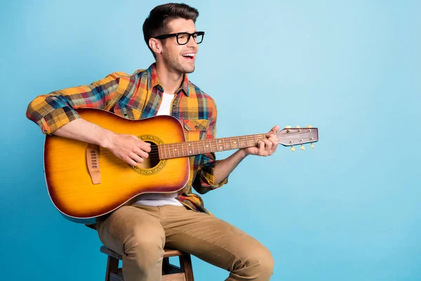 Foto de cara muito adorável vestido xadrez camisa óculos sorrindo sentado cadeira tocando guitarra espaço vazio isolado azul cor de fundo — Fotografia de Stock