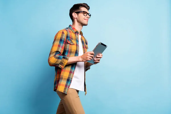 Фото смешного милого парня, одетого в клетчатую рубашку, улыбающегося, держащего современный гаджет, идущего, глядя на пустое пространство изолированного синего цвета фона — стоковое фото