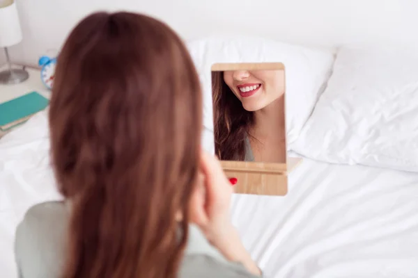 Achterzijde achteraanzicht portret van aantrekkelijk vrolijk meisje zittend op bed kijkend naar spiegelreflexie thuis huis licht wit interieur — Stockfoto