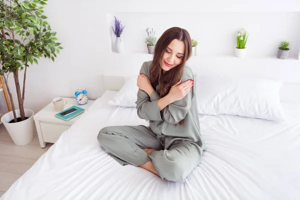Фото очаровательной молодой женщины в серой ночной рубашке, сидящей на кровати и обнимающей себя, улыбающейся в помещении. — стоковое фото