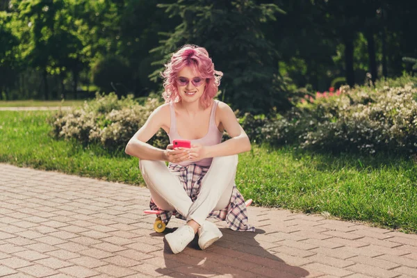 Foto in voller Länge von jungen fröhlichen Mädchen glücklich positives Lächeln sitzen Longboard-Look suchen Handy im Freien — Stockfoto