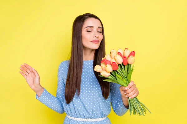 Mavi elbiseli, sevimli genç bir kadının fotoğrafı. Çiçek aromasının tadını çıkarıyor. Gülümsüyor, izole edilmiş sarı arka plan. — Stok fotoğraf