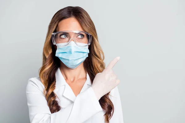Düşünceli, kendine güvenen, beyaz önlüklü, gözlüklü, boş bir yeri işaret eden gri arkaplanı olan bir kadın doktor fotoğrafı. — Stok fotoğraf