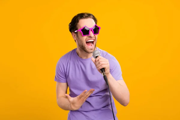 Fénykép őrült szupersztár énekes srác tartja mikrofon sing viselet napszemüveg lila póló izolált sárga színű háttér — Stock Fotó