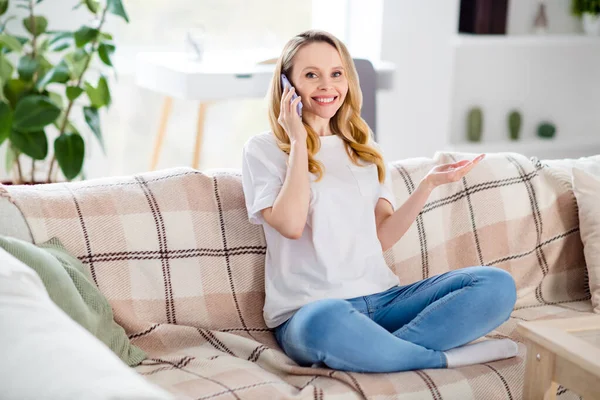 Фотография в полный рост молодой привлекательной женщины, счастливой позитивной улыбкой сидящей на диване и говорящей по мобильному телефону самоизоляции карантина в помещении — стоковое фото