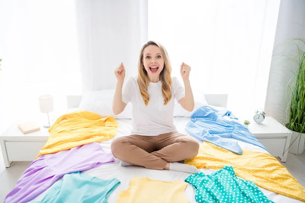 Retrato de chica alegre atractiva sentada en la cama que se divierte eligiendo la ropa en la casa de luz blanca en el interior —  Fotos de Stock