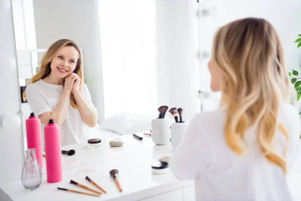 Retrato de chica rubia alegre atractiva mirando el espejo disfrutando del procedimiento de maquillaje de descanso matutino en el hogar blanco claro en el interior — Foto de Stock