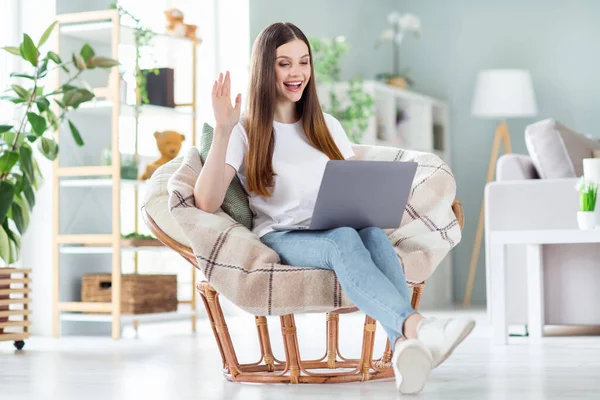 Retrato de chica alegre atractiva sentada en silla usando el ordenador portátil llamando saludando en casa sala de estar de luz en el interior —  Fotos de Stock