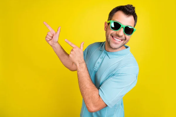 Foto di funky grazioso giovane ragazzo vestito blu t-shirt occhiali scuri sorridente indicando spazio vuoto isolato colore giallo sfondo — Foto Stock