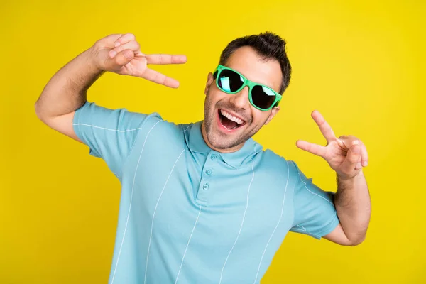 Foto van funky brunet man show v-teken slijtage bril blauw t-shirt geïsoleerd op gele kleur achtergrond — Stockfoto