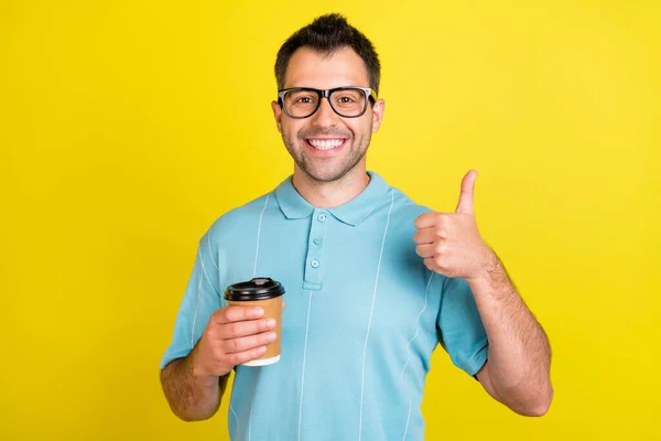 Foto de la divertida morena hombre sostener café mostrar pulgar hacia arriba usar gafas azul camiseta aislada sobre fondo de color amarillo —  Fotos de Stock