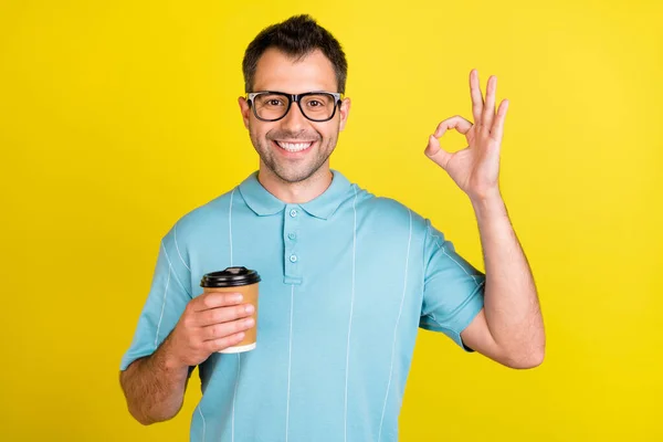 Foto do homem morena legal segurar café mostrar okey sinal desgaste óculos azul t-shirt isolado no fundo de cor amarela — Fotografia de Stock