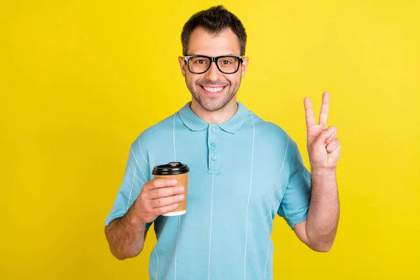 Foto de engraçado morena homem segurar café show v-sign desgaste óculos azul t-shirt isolado no fundo de cor amarela — Fotografia de Stock