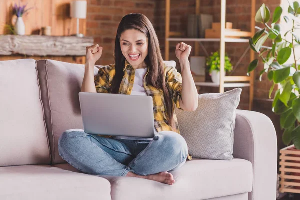 Foto wanita tercengang duduk nyaman sofa memegang netbook mengangkat kepalan tangan merayakan kemenangan mengenakan kemeja kotak-kotak rumah tangga — Stok Foto