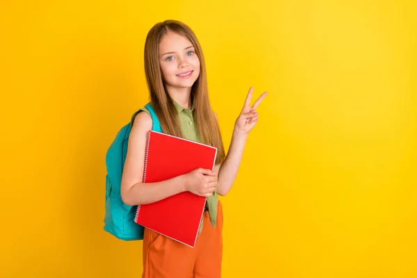 Foto retrato colegiala vistiendo camisa verde mochila mantenimiento libro pila mostrando v-signo aislado vívido color amarillo fondo — Foto de Stock