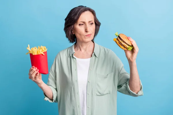 Foto porträtt senior kvinna äter stekt potatis burgare osäker fick tvivel isolerad pastell blå färg bakgrund — Stockfoto