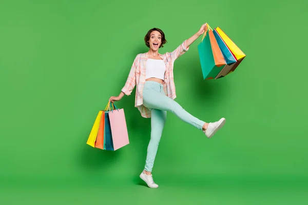 Foto de cuerpo completo de alegre mujer joven feliz levantar las manos pierna mantenga muchos paquetes comprador aislado sobre fondo de color verde —  Fotos de Stock