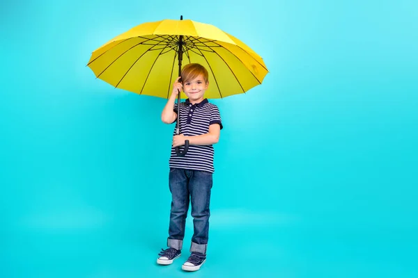 İyimser küçük çocuğun tam vücut fotoğrafı şemsiye tut, tişört giy, spor ayakkabılarını deniz mavisi arka planına izole et. — Stok fotoğraf