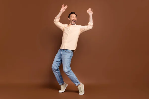 Zdjęcie beztroski marzycielski facet taniec podnieść ręce stać paluszki nosić beżowy koszula dżinsy buty odizolowany brązowy kolor tło — Zdjęcie stockowe
