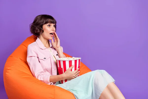 젊은 여성 의자에 앉아 스낵을 먹는 사진 보라색 배경에 고립된 빈 공간 TV 를 보고 놀라는 여성의 사진 — 스톡 사진