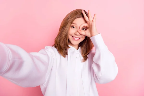 Fénykép édes fényes iskolás lány viselni fehér kapucnis mosolygós tacking selfie okey jel fedél szem elszigetelt rózsaszín szín háttér — Stock Fotó