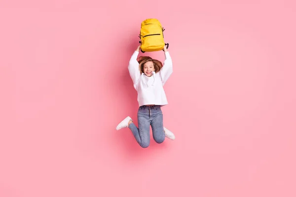 En jente på full lengde som hopper høyt, holder skolesekken over hodet, isolert, pastellrosa, farget bakgrunn. – stockfoto