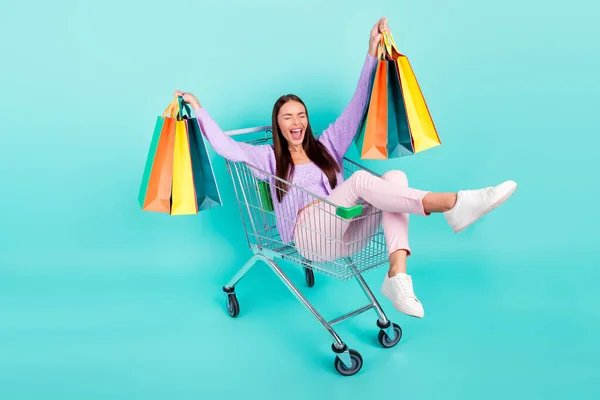 Foto de dulce mujer funky vestida ropa púrpura sosteniendo bolsas montando bandeja de compras riendo sonriente color verde azulado aislado fondo — Foto de Stock