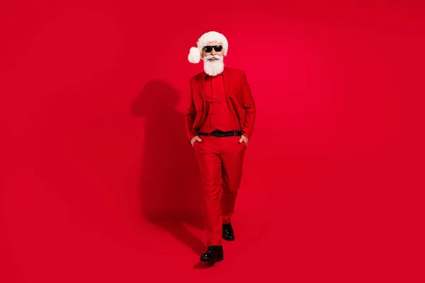 Foto de confianza guapo santa claus manos bolsillos usar gafas de sol x-mas sombrero traje zapatos sobre fondo de color rojo —  Fotos de Stock