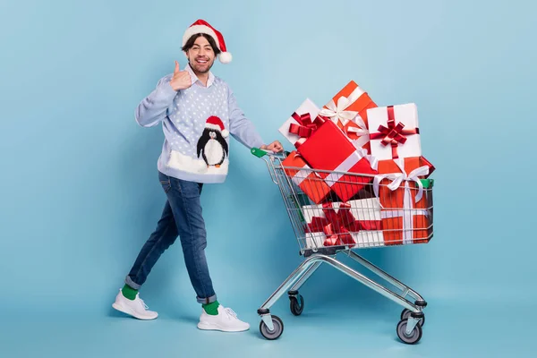 Ganzkörperfoto von brünetten Mann machen Daumen nach oben Warenkorb Geschenke tragen Jeans Weihnachtspullover Weihnachtsmann Hut isoliert auf blauem Hintergrund — Stockfoto