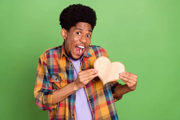 Foto de chico de piel oscura dulce impresionado vestido camisa a cuadros sosteniendo corazón de madera sonriendo aislado color verde fondo — Foto de Stock