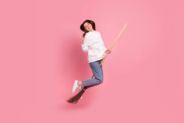 フルボディプロフィール写真の若いです女性ジャンプオンほうき着用パーカージーンズスニーカー隔離されたピンクの背景 — ストック写真
